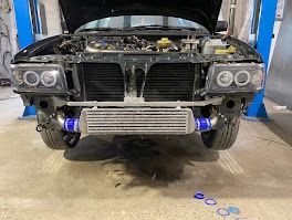 Czarny samochód z otwartą maską i zamontowanym intercoolerem, widoczne nowe reflektory z ringami. Warsztatowy podnośnik w tle, ślady opon na posadzce.