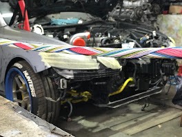 Przód szarego auta w warsztacie, z widocznym turbodoładowaniem i elementami tuningowymi. Taśma zabezpieczająca na karoserii, niebieskie felgi. Silnik w tle.