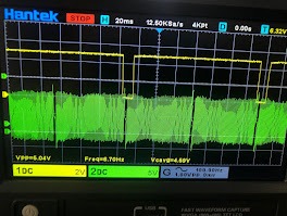 Oscyloskop Hantek z wyświetlonymi przebiegami sygnałów elektrycznych w kolorach żółtym i zielonym. Widoczne parametry pomiarowe na ekranie urządzenia.