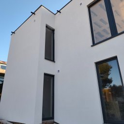 Nowoczesny, dwukondygnacyjny budynek z jasną elewacją i czarnymi ramami okiennymi. Widoczne detale wykończenia i fragment terenu przed budynkiem.