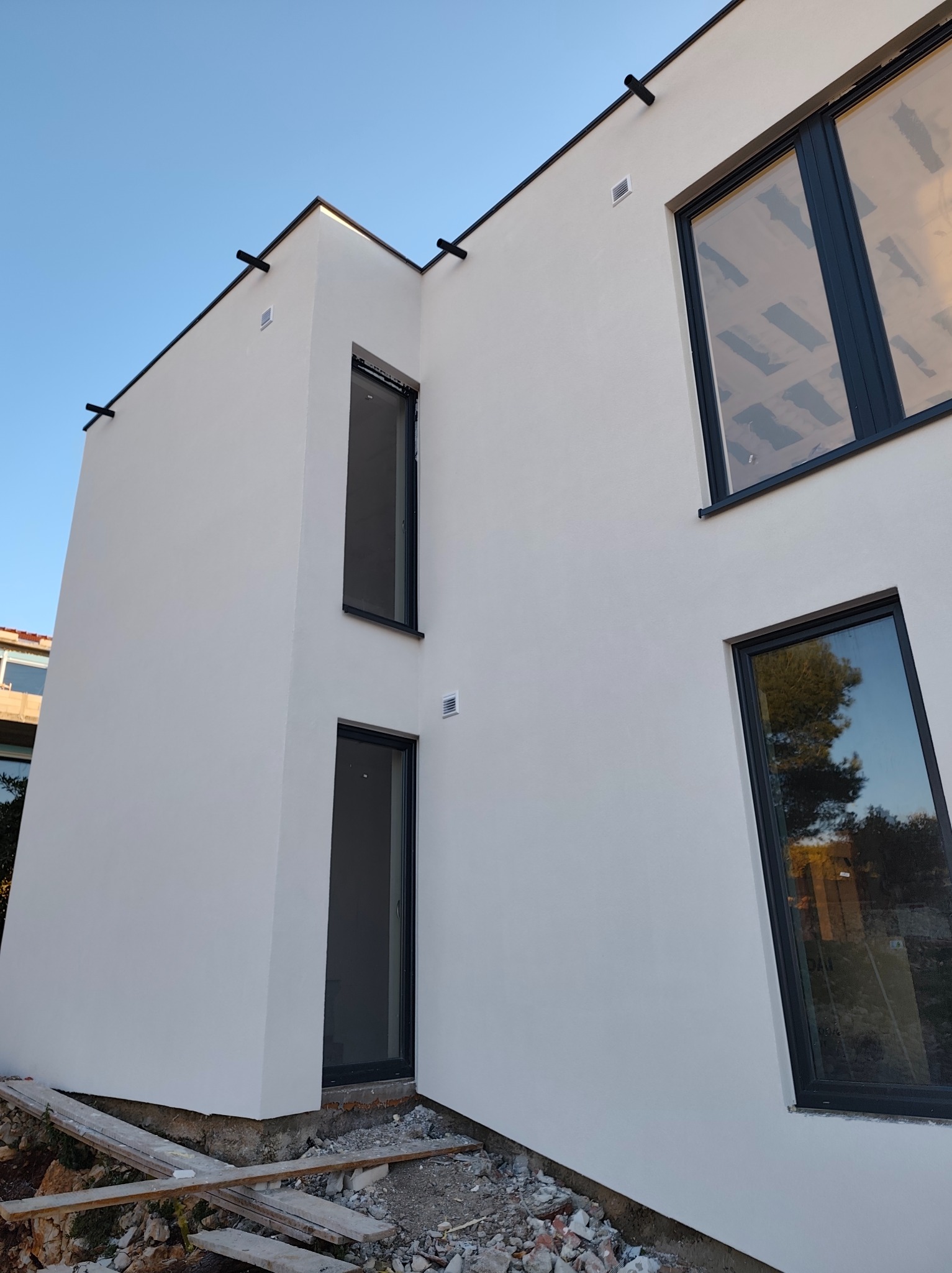 Nowoczesny, dwukondygnacyjny budynek z jasną elewacją i czarnymi ramami okiennymi. Widoczne detale wykończenia i fragment terenu przed budynkiem.