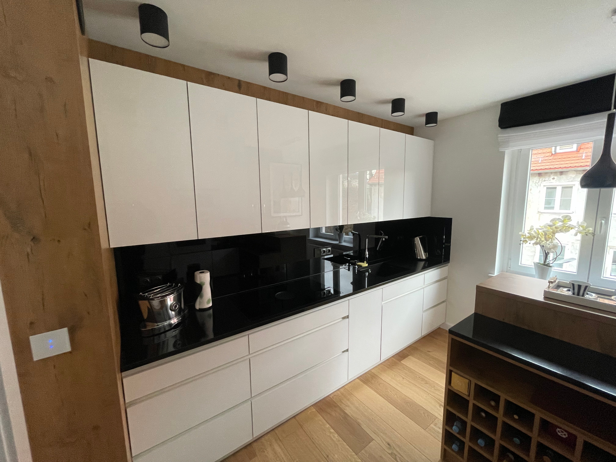 Nowoczesna kuchnia z białymi, lakierowanymi frontami, czarnym blatem i oświetleniem punktowym. Widoczny fragment drewnianej zabudowy i okno z widokiem na zewnątrz.