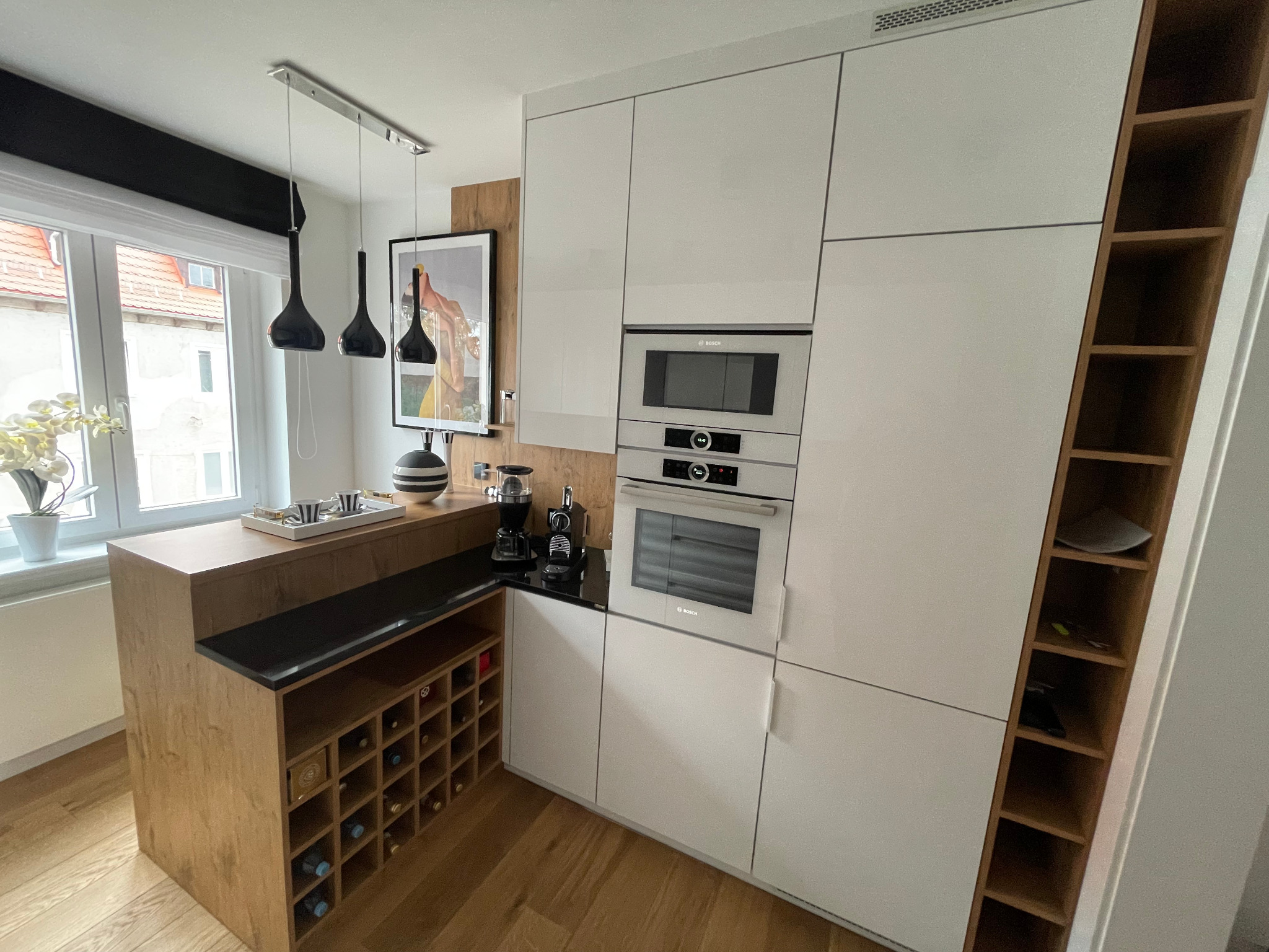 Nowoczesna kuchnia z białymi szafkami, drewnianym blatem i półkami na wino. Widoczne sprzęty AGD marki Bosch i designerskie lampy wiszące.