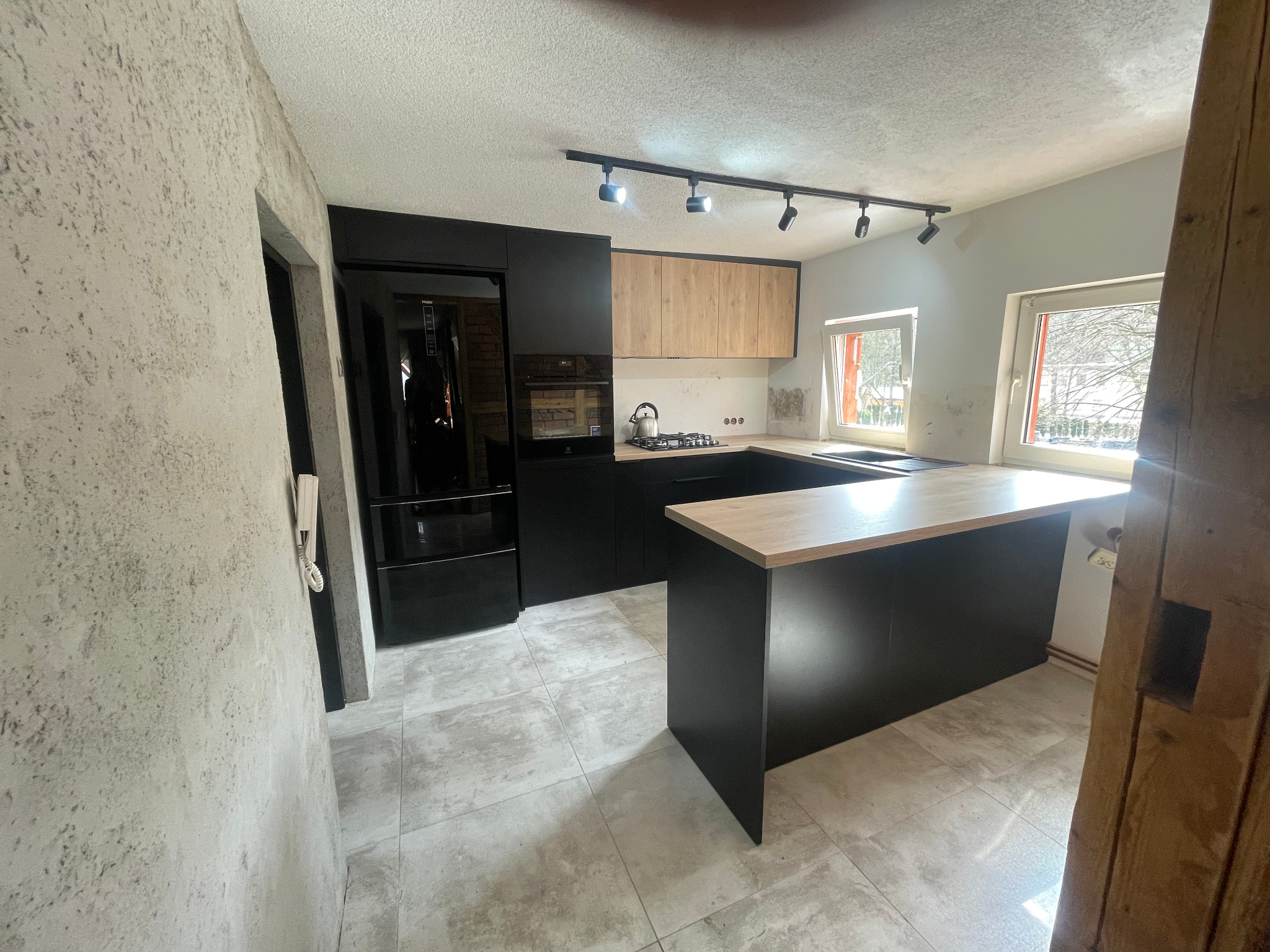 Nowoczesna kuchnia z czarnymi szafkami i drewnianym blatem, oświetlona listwą LED. Widok na wyspę kuchenną i sprzęt AGD w zabudowie. Minimalistyczny design.