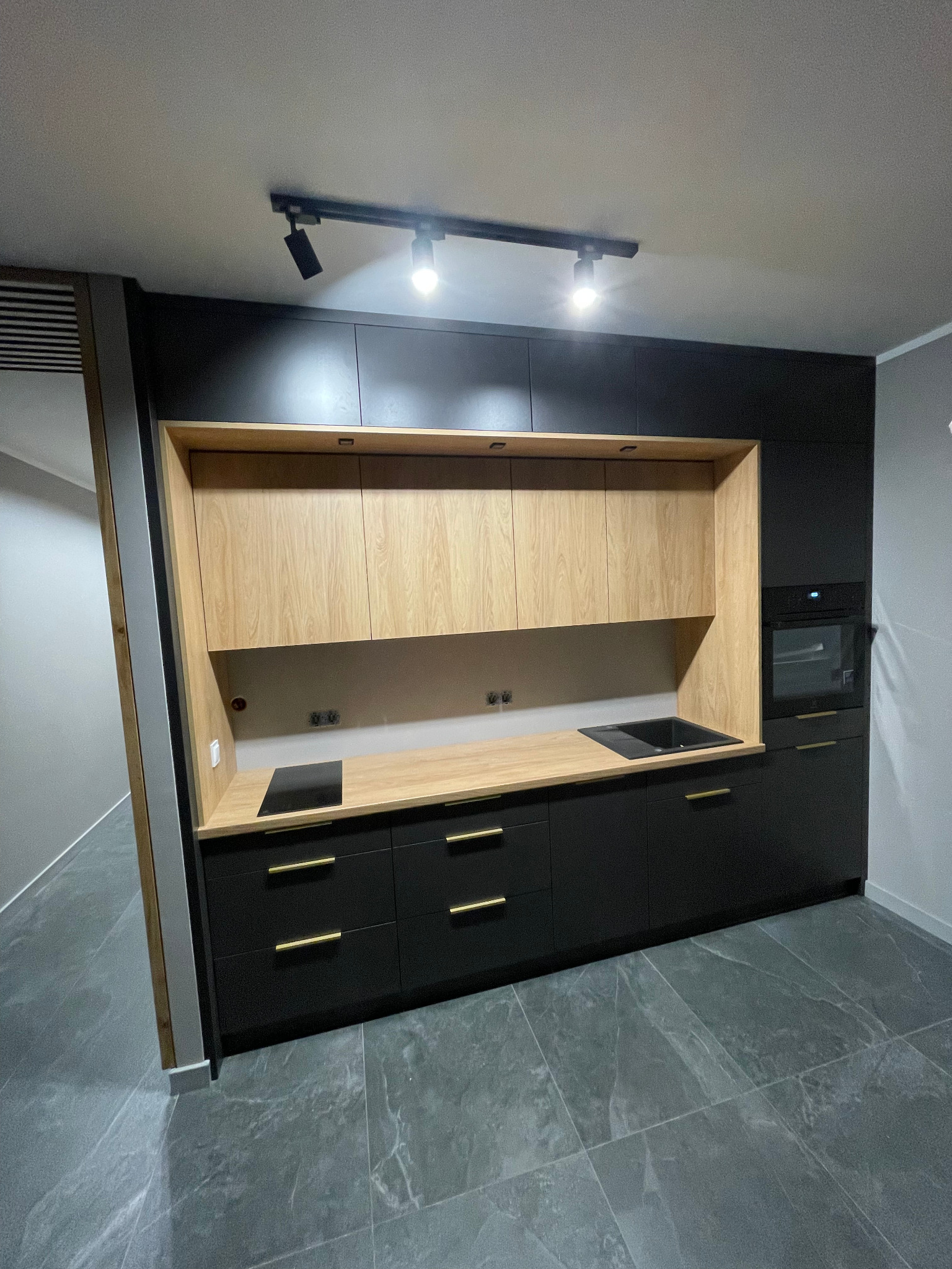 Nowoczesna kuchnia na wymiar: czarne szafki z drewnianym blatem i złotymi uchwytami, oświetlenie punktowe, szare płytki podłogowe. Minimalistyczny design.