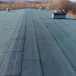 R-dach Robert Szelwach - Niebieski dach pokryty papą termozgrzewalną z kominem i odpowietrznikami na tle pagórkowatego krajobrazu w słoneczny dzień. Widok z góry.