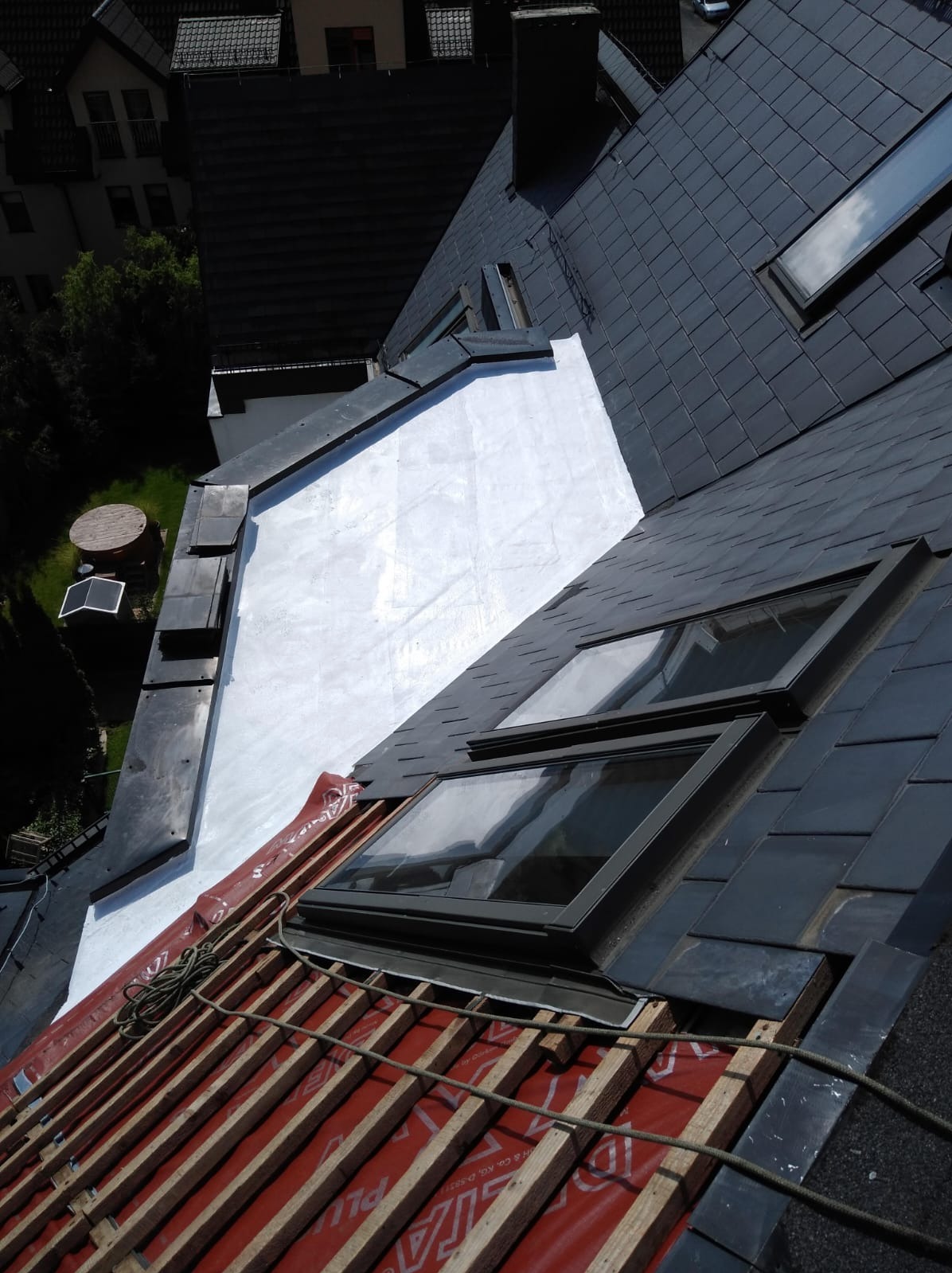 Częściowy widok dachu z czarną dachówką, oknami połaciowymi i fragmentem konstrukcji z czerwoną membraną. Biała, gładka powierzchnia przylega do dachu.