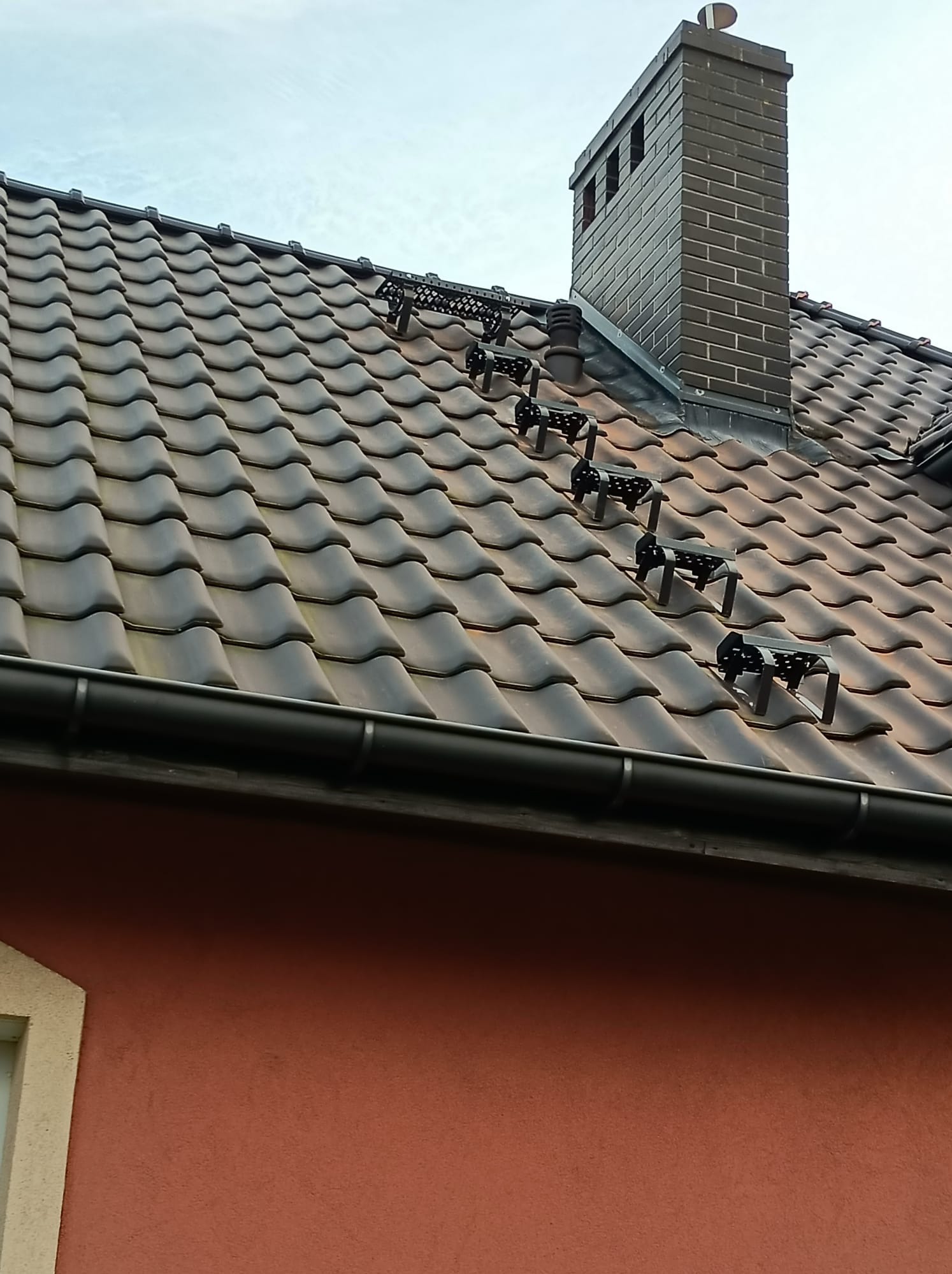 Brązowy dach z dachówki ceramicznej z zamontowanymi stopniami kominiarskimi i ceglanym kominem. Widoczna rynna i fragment ściany budynku.