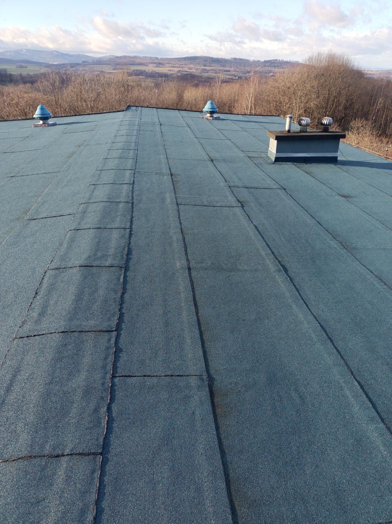 Niebieski dach pokryty papą termozgrzewalną z kominem i odpowietrznikami na tle pagórkowatego krajobrazu w słoneczny dzień. Widok z góry.