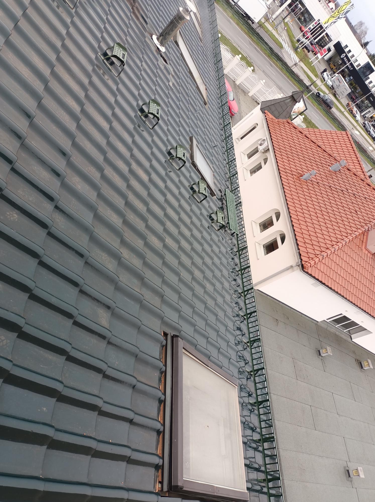 Ciemny dach z metalowej dachówki z oknami dachowymi i uchwytami przeciwśniegowymi. Widoczna drabina dachowa i fragment sąsiedniego budynku z czerwoną dachówką.