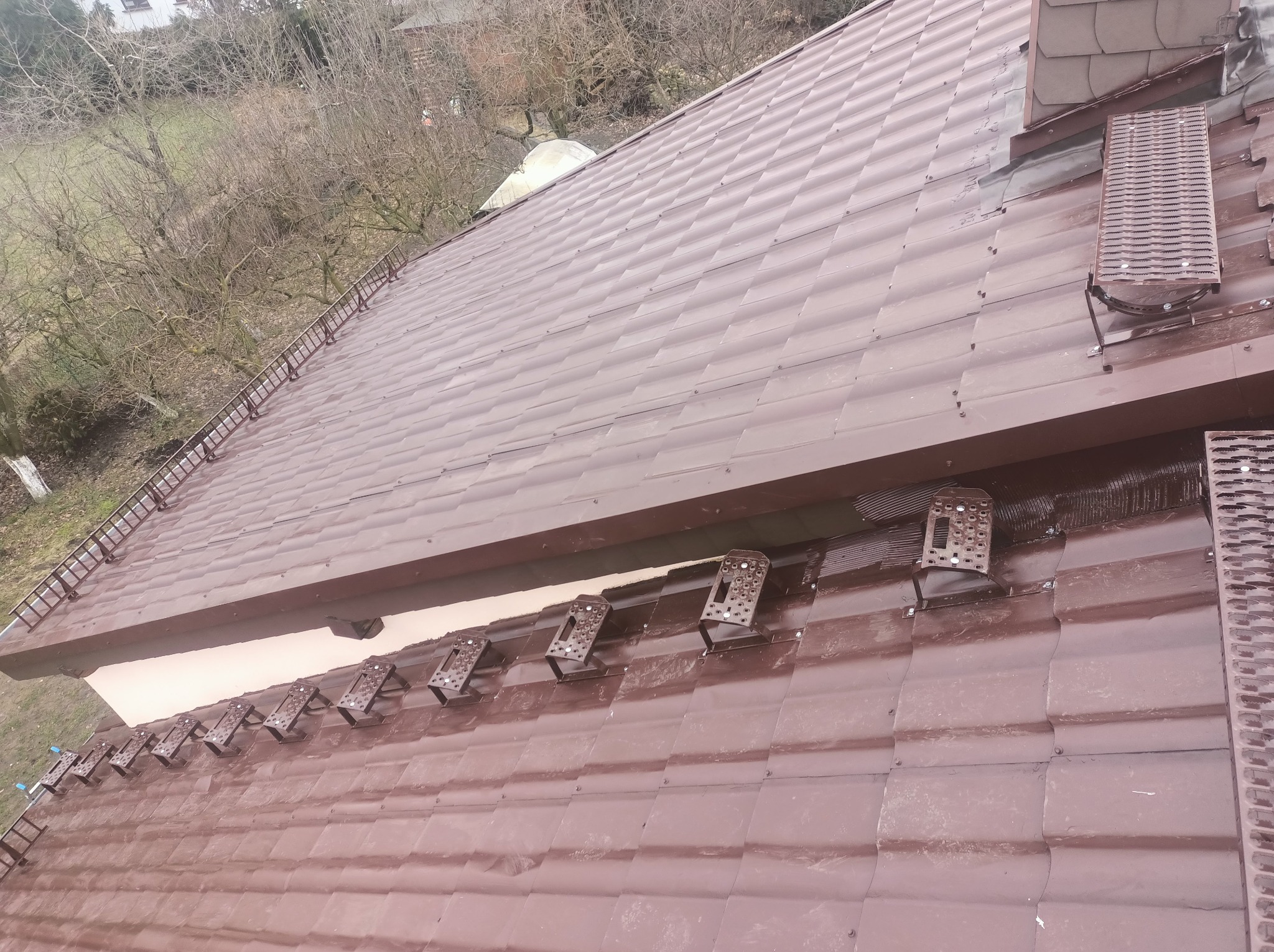 Ciemnobrązowy dach z zamontowanymi stopniami kominiarskimi i drabinką dachową, widok z góry na tle drzew bez liści.