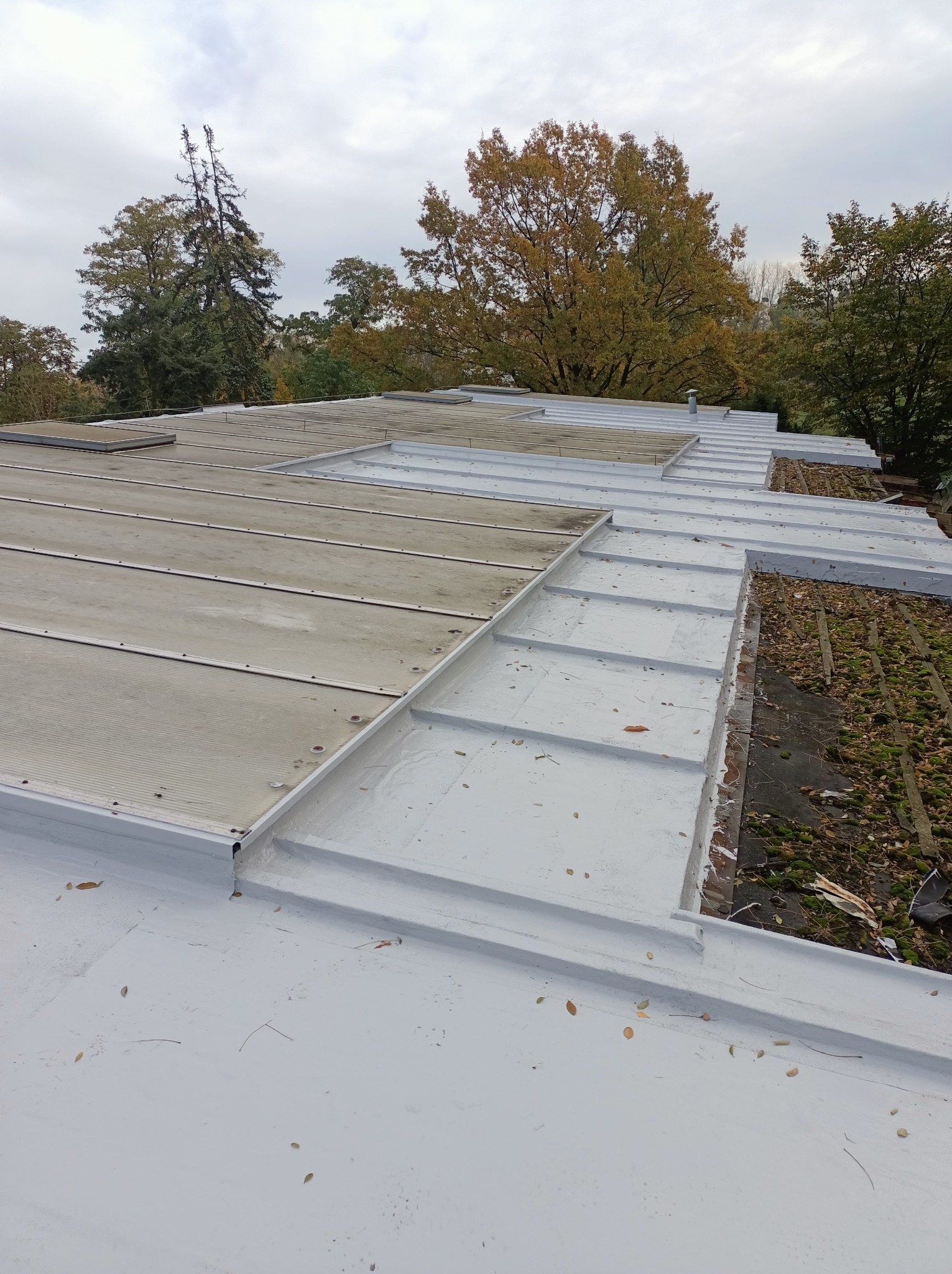 Biały dach z panelami i elementami blacharskimi w trakcie prac. Widoczne fragmenty starego pokrycia i otoczenie drzew. Pochmurne niebo w tle.