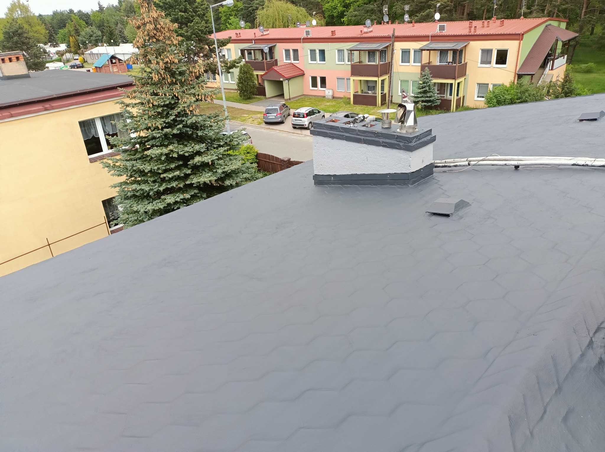 Szary dach z kominem na tle osiedla mieszkaniowego i zieleni. Widoczne kable na dachu. Dach pokryty papą w heksagonalny wzór. Ujęcie z góry, lekko z ukosa.
