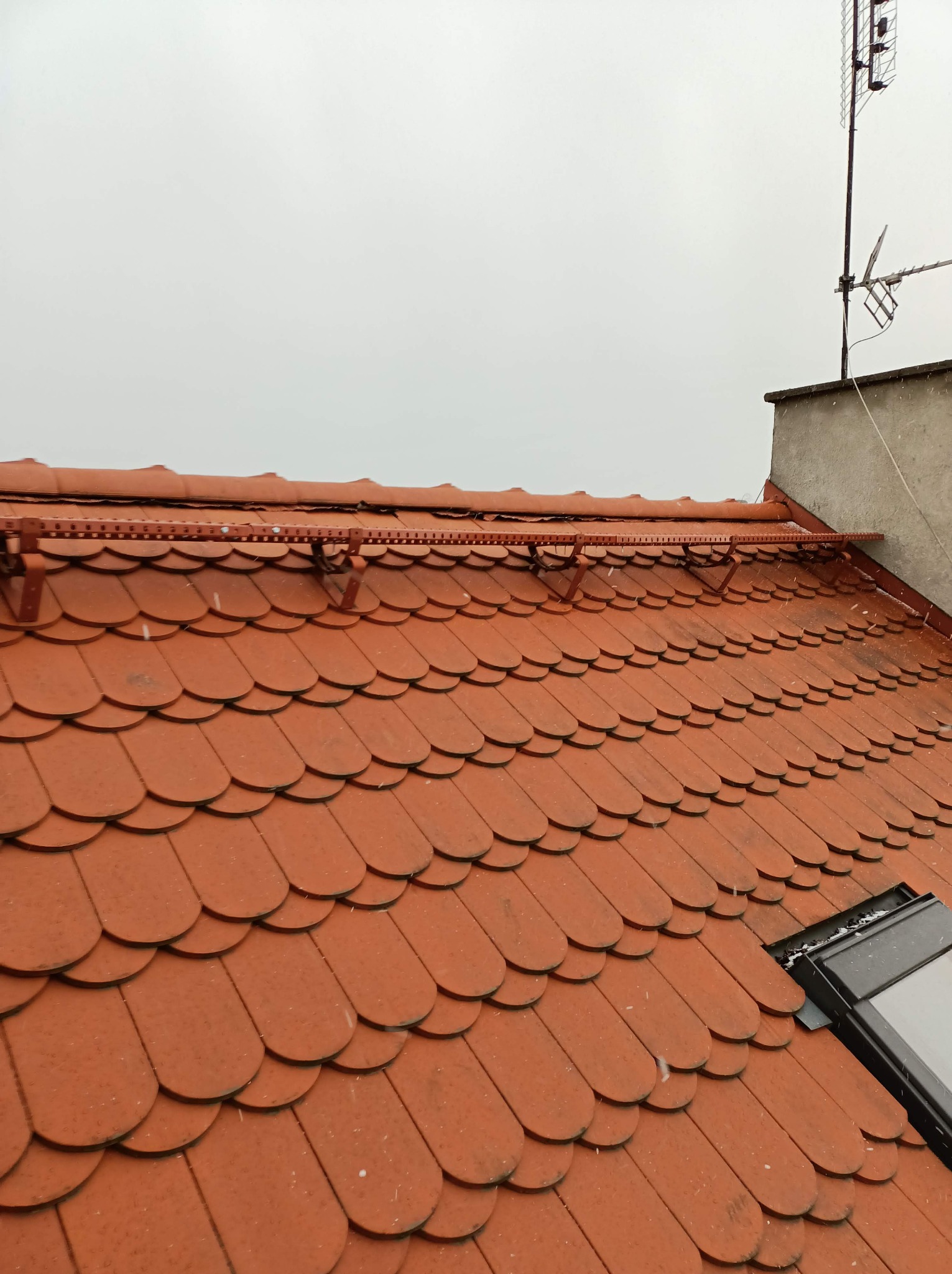 Czerwony dach pokryty dachówką karpiówką z zamontowanym systemem przeciwśniegowym i oknem dachowym, widok z góry w pochmurny dzień.