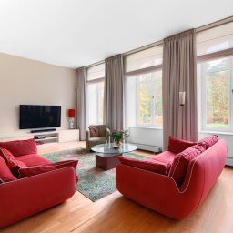 Klaudia Walter-Jurgasz - Jasny salon z parkietem, czerwonymi sofami, dywanem i widokiem na zieleń za oknem. Nowoczesny design z telewizorem i dekoracjami.