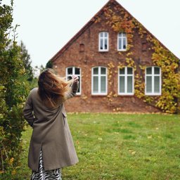 Klaudia Walter-Jurgasz - Kobieta w płaszczu stoi tyłem na trawie, w tle ceglany dom z pnączami. Ujęcie z dołu, podkreślające wysokość budynku i jesienny charakter otoczenia.