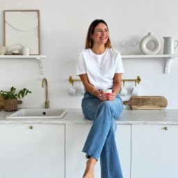 Klaudia Walter-Jurgasz - Uśmiechnięta kobieta siedzi na blacie w jasnej kuchni z marmurowym blatem, trzymając kubek. Minimalistyczny wystrój, złota bateria i drewniana deska do krojenia.
