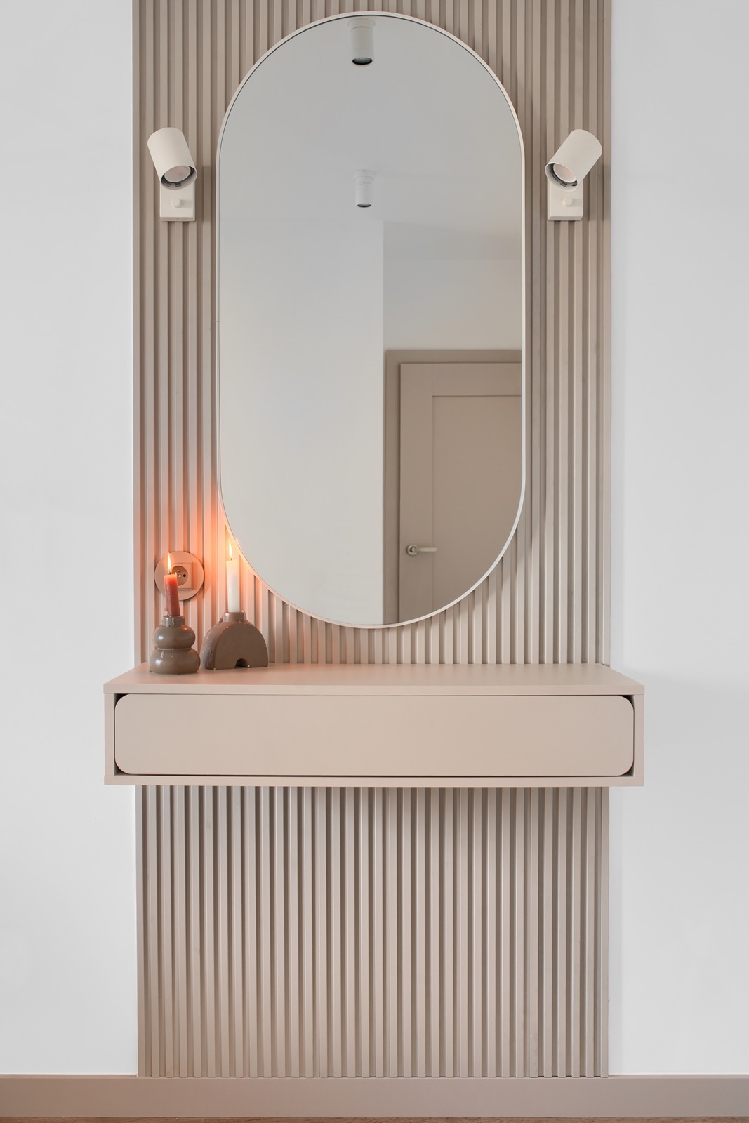 Beżowa konsola z owalnym lustrem i świecami na tle ściany z pionowych paneli. Minimalistyczny design, ciepłe oświetlenie.