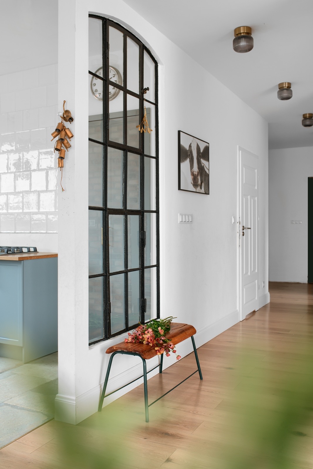 Jasne wnętrze z drewnianą podłogą, ławką i kwiatami przy oknie w stylu industrialnym. W tle widoczna kuchnia z niebieskimi szafkami i białe ściany z dekoracjami.