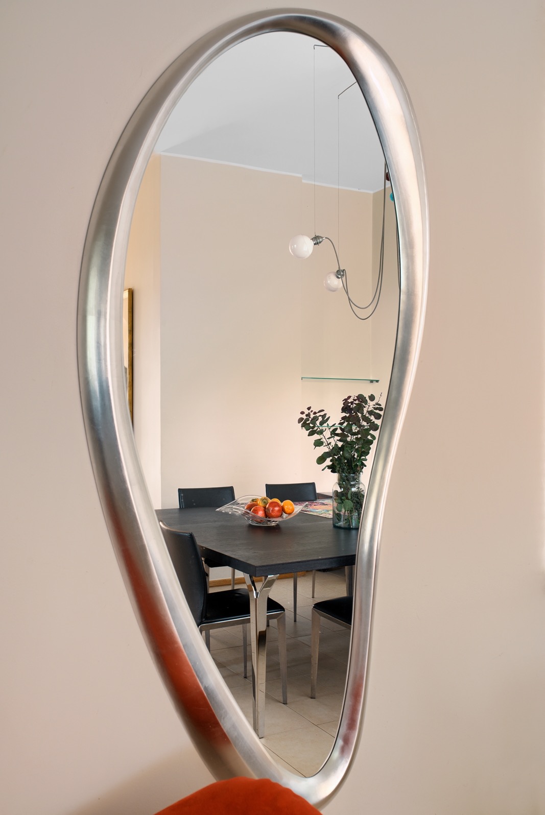 Nietypowe lustro w srebrnej ramie odbija wnętrze jadalni: czarny stół z owocami, krzesła i wiszące lampy. Minimalistyczny design, ciepłe oświetlenie.
