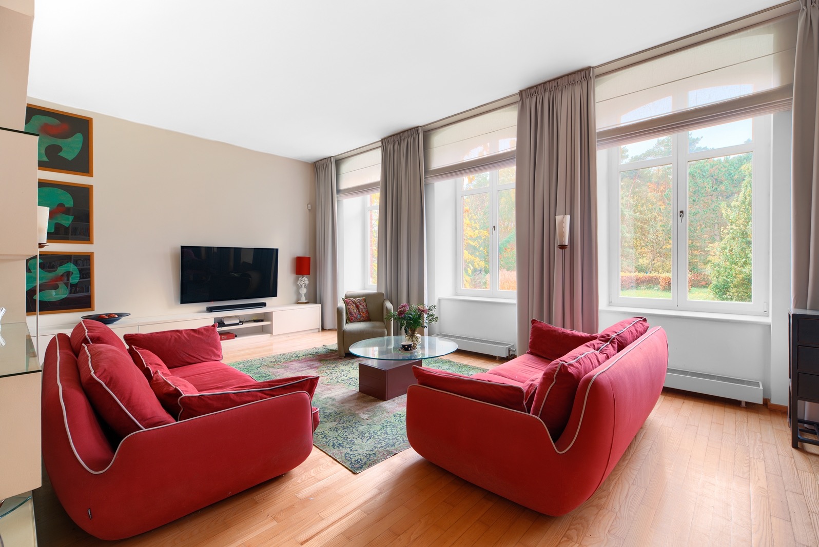 Jasny salon z parkietem, czerwonymi sofami, dywanem i widokiem na zieleń za oknem. Nowoczesny design z telewizorem i dekoracjami.