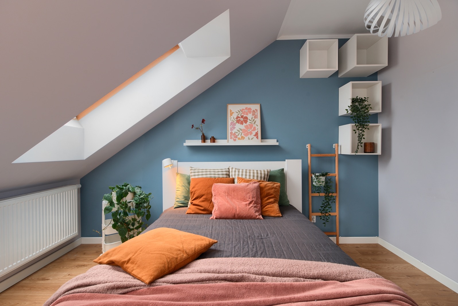 Przytulna sypialnia na poddaszu z łóżkiem, dekoracjami i oknem dachowym. Niebieska ściana, drewniana podłoga i białe półki tworzą ciepłą atmosferę.