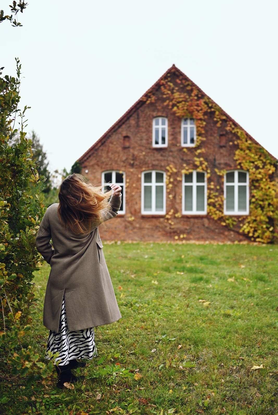 Kobieta w płaszczu stoi tyłem na trawie, w tle ceglany dom z pnączami. Ujęcie z dołu, podkreślające wysokość budynku i jesienny charakter otoczenia.