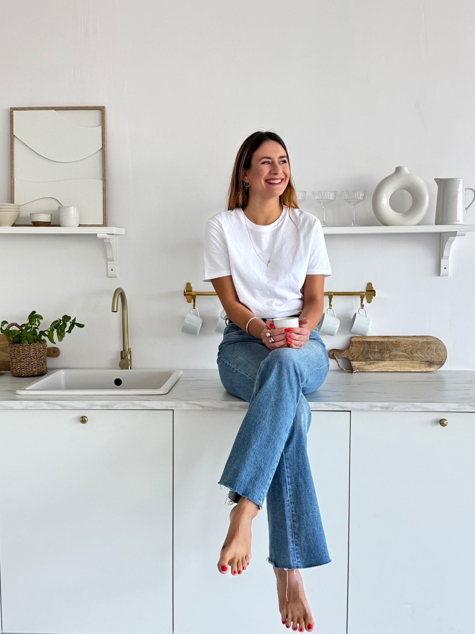 Uśmiechnięta kobieta siedzi na blacie w jasnej kuchni z marmurowym blatem, trzymając kubek. Minimalistyczny wystrój, złota bateria i drewniana deska do krojenia.