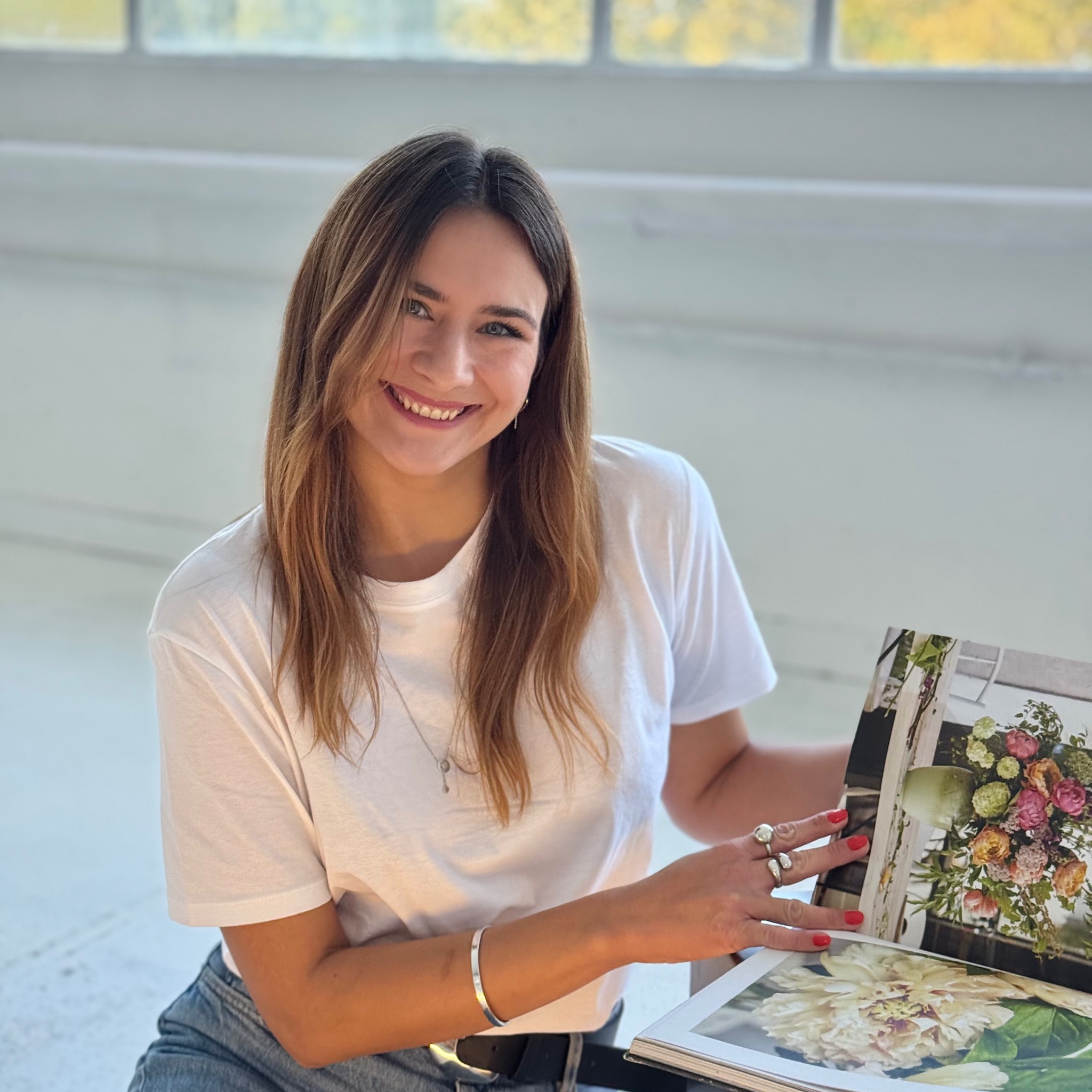 Uśmiechnięta kobieta w białym t-shircie przegląda album z fotografiami kwiatów w jasnym wnętrzu. Widoczne detale: srebrna biżuteria i czerwony manicure.
