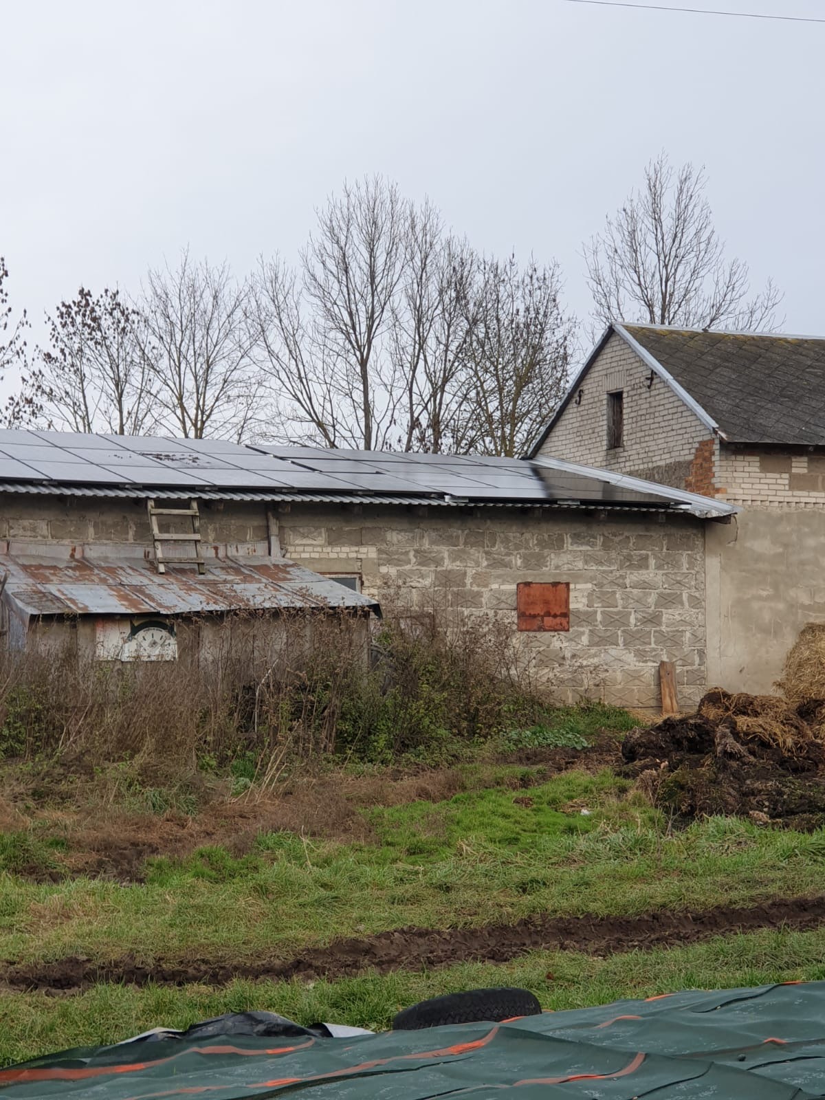 Instalacja fotowoltaiczna 18 kWp na dachu budynku gospodarczego z cegły, widoczna drabina oparta o dach, pochmurne niebo w tle.