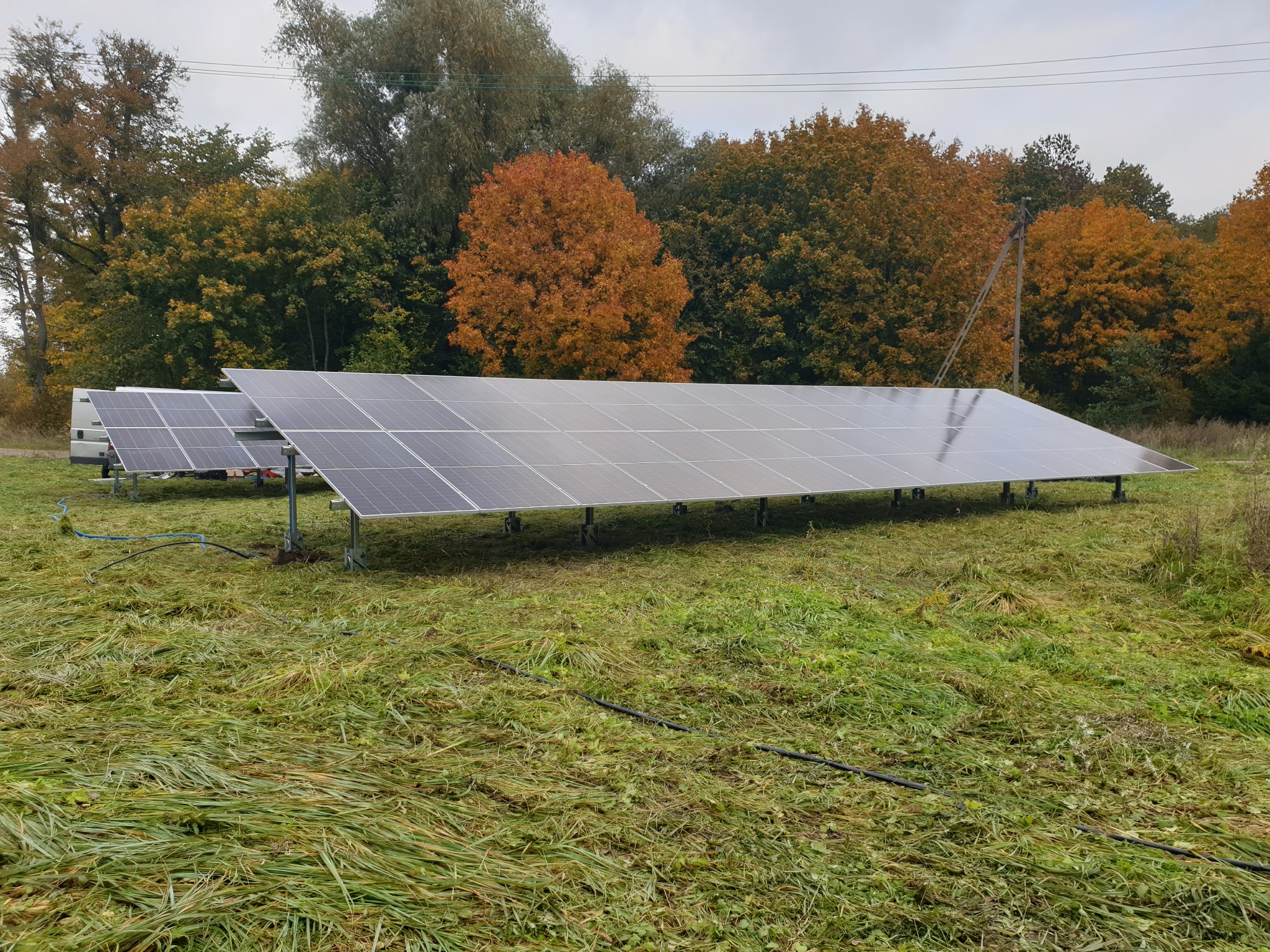Panel fotowoltaiczny 30 kWp na łące, jesienne drzewa w tle. Widoczne kable i konstrukcja wsporcza. Ujęcie z poziomu gruntu, pochmurne niebo.