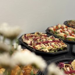 SOSNOWIECKA SIEĆ SZEROKOPASMOWA SPÓŁKA Z OGRANICZONĄ ODPOWIEDZIALNOŚCIĄ - Elegancko ułożone wrapy z warzywami i mięsem na czarnych tacach, część kompozycji zasłonięta przez białe kwiaty. Catering na czarnym stole, białe tło.