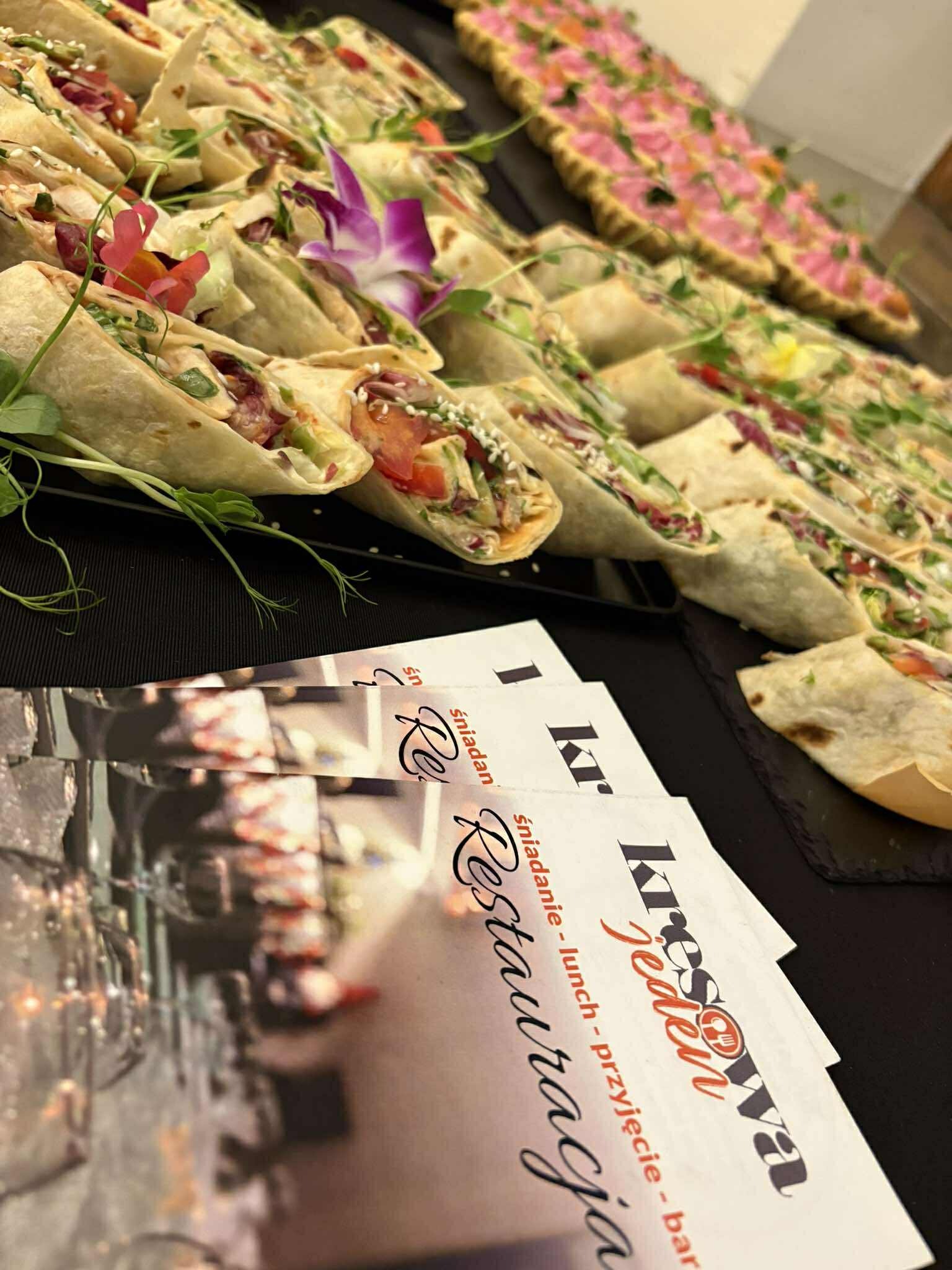 Catering: Rządki wrapów z warzywami i mięsem, dekorowane kwiatami i zieleniną, obok ulotki restauracji. Elegancki bufet na przyjęcie.