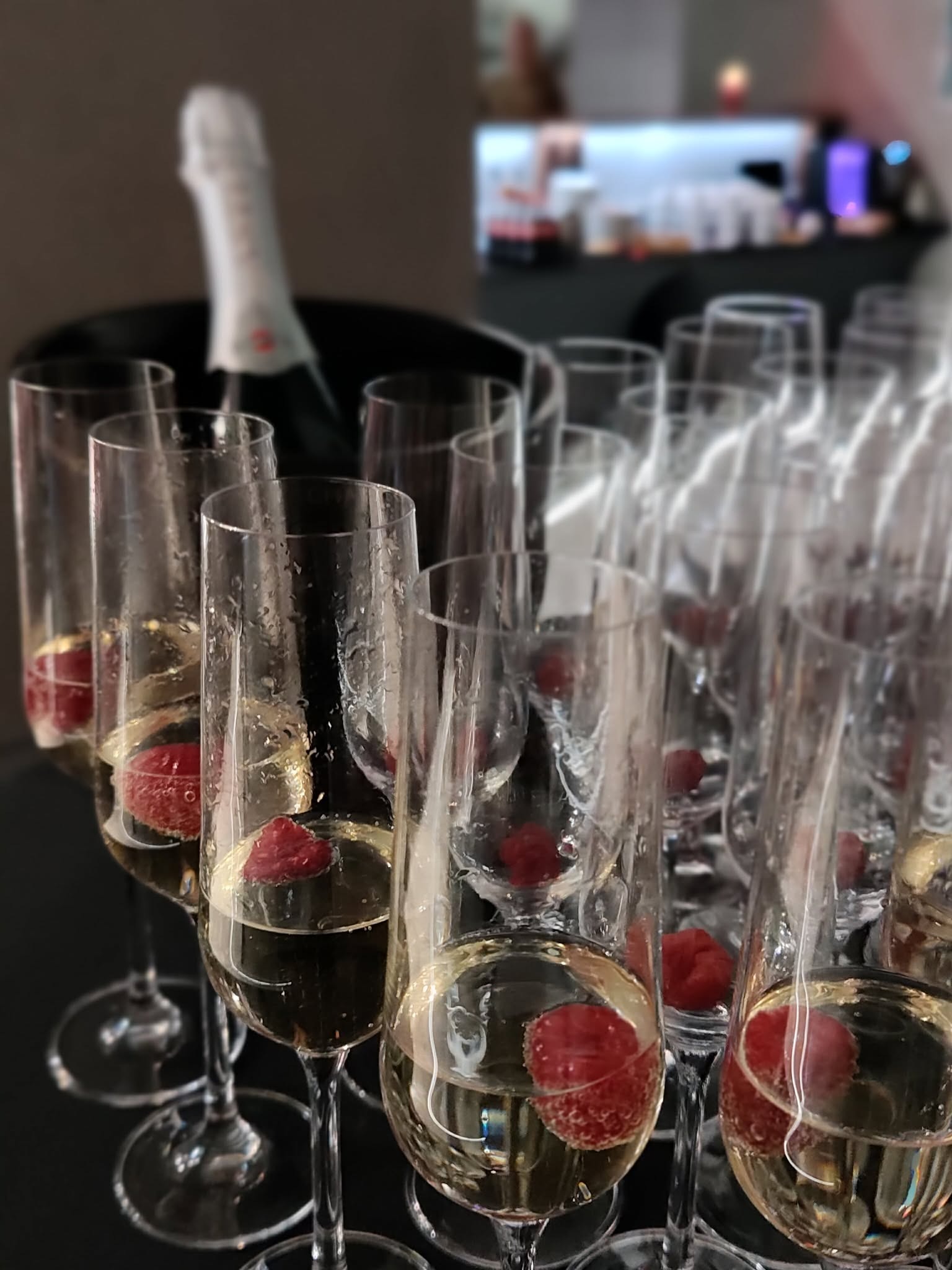 Szampan z malinami w smukłych kieliszkach na czarnej tacy, butelka w tle. Elegancki catering na przyjęciu, delikatne światło.