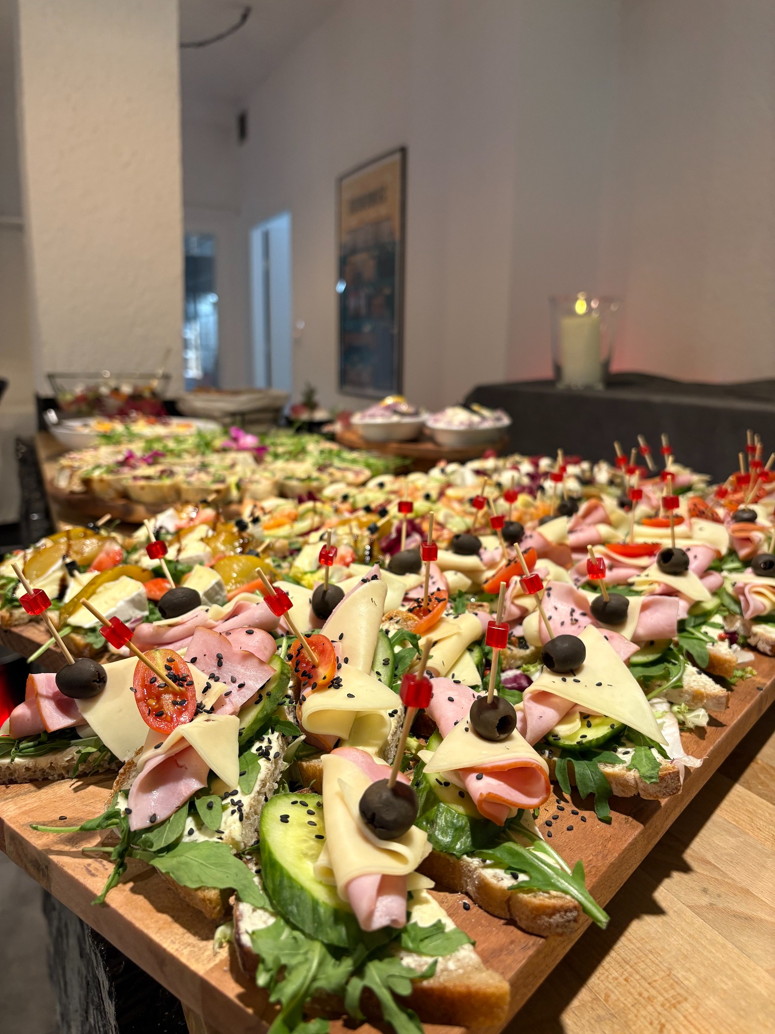 Bogato zdobione kanapki na drewnianej desce, idealne na event w Sosnowcu. Różnorodność składników: szynka, ser, warzywa, oliwki. Elegancki catering.