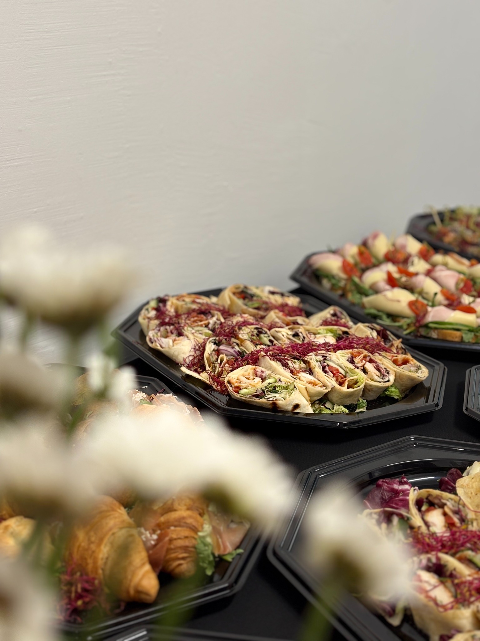 Elegancko ułożone wrapy z warzywami i mięsem na czarnych tacach, część kompozycji zasłonięta przez białe kwiaty. Catering na czarnym stole, białe tło.