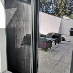 PergoleTarasy.pl - Zasłonięta plisowana moskitiera w szarej ramie, zamontowana na wyjściu na taras. Widoczny fragment tarasu z meblami ogrodowymi i drzewo w tle.