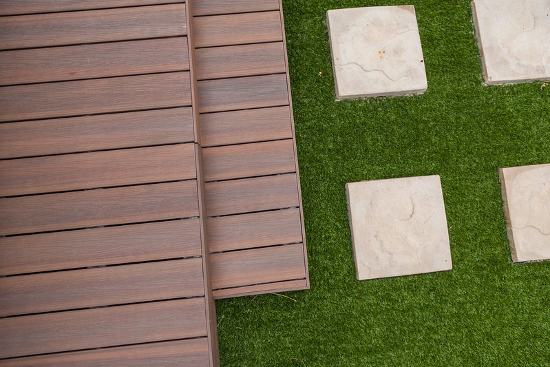 Kompozytowy taras w odcieniach brązu, z elementami schodów i ścieżką z kwadratowych płyt na sztucznej trawie. Widok z góry, podkreślający strukturę desek.