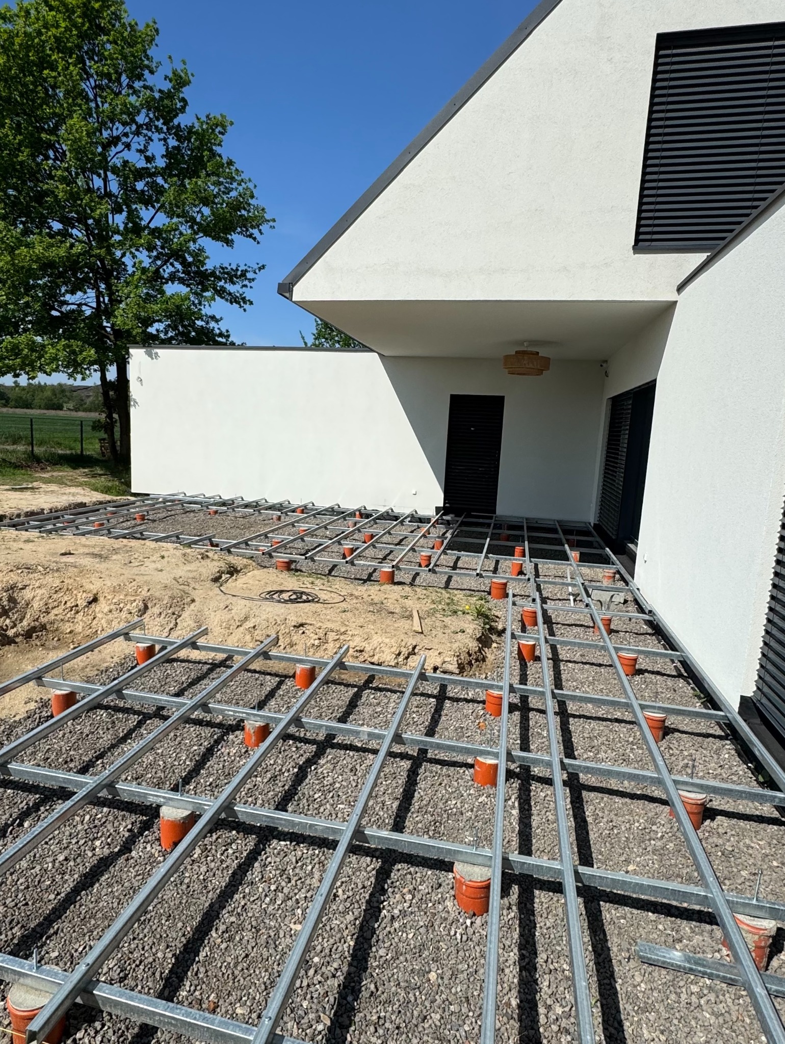 Montaż metalowej konstrukcji tarasu na żwirowym podłożu, widoczne pomarańczowe wsporniki. Nowoczesny dom z białymi ścianami i czarnymi oknami w tle, błękitne niebo.