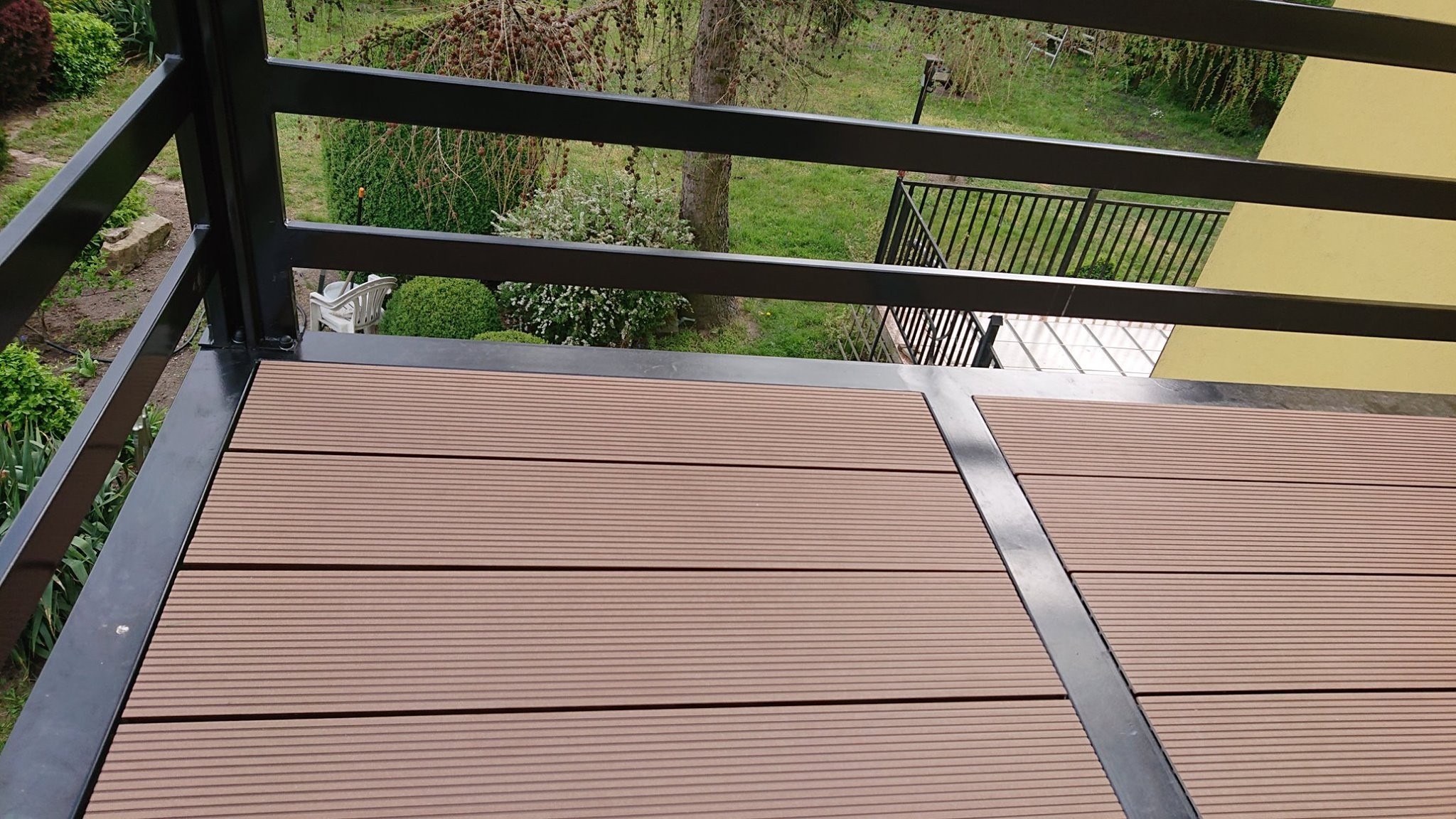 Nowoczesny taras kompozytowy z czarną metalową balustradą, widok z góry na deski w odcieniach brązu i otaczającą zieleń ogrodu.