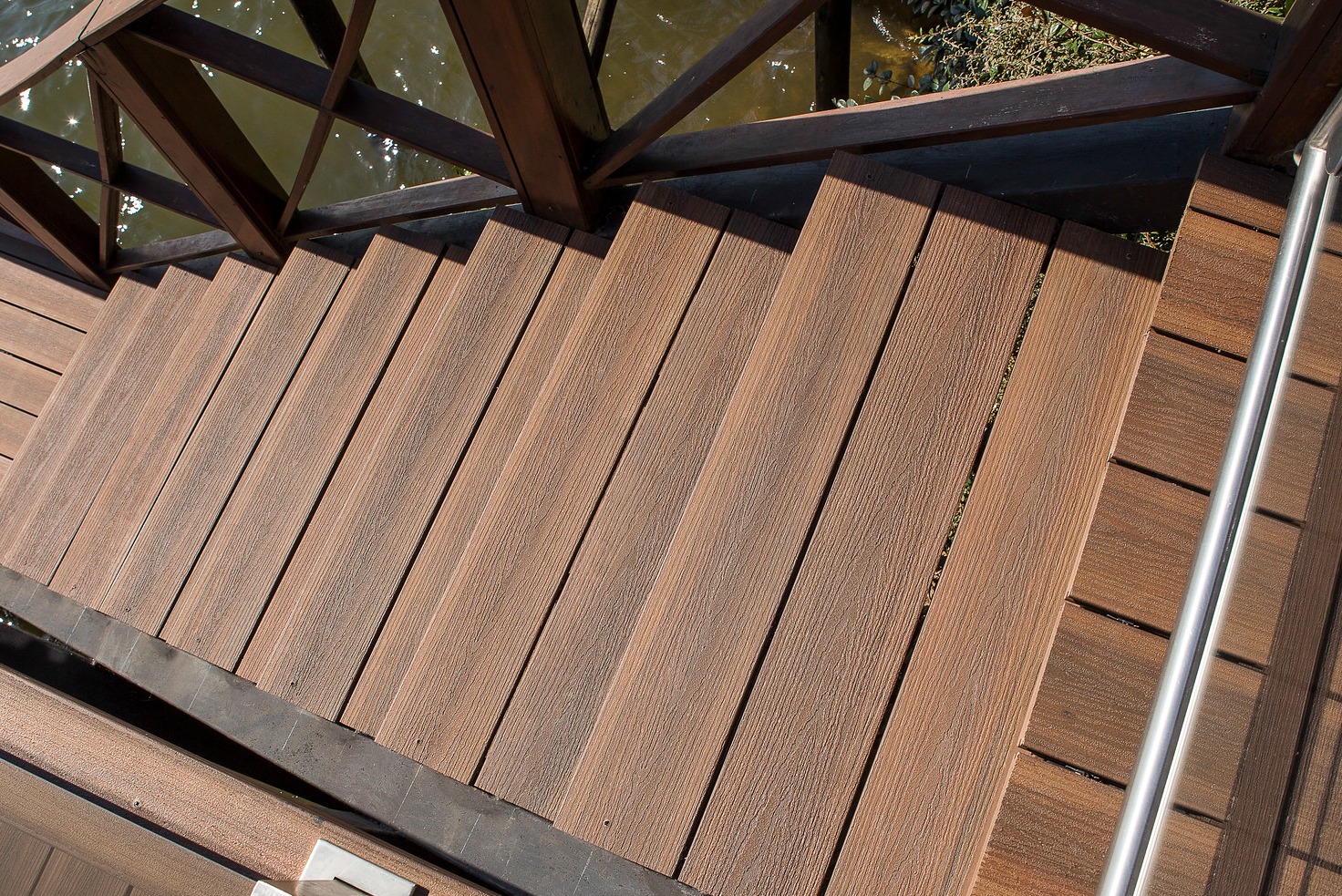 Schody z desek kompozytowych prowadzące do wody, z drewnianą balustradą i metalową poręczą. Widok z góry, ukazujący strukturę desek i otoczenie wodne.