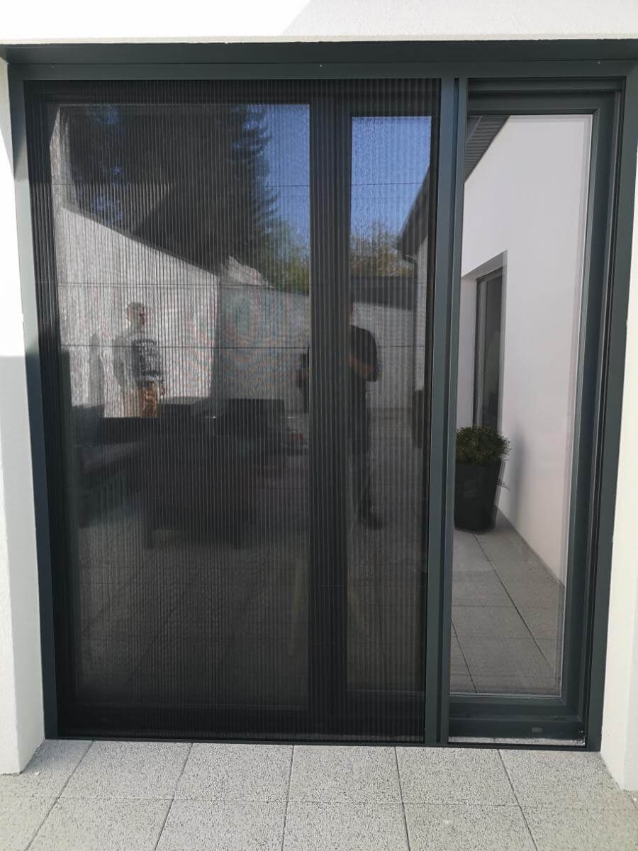 Zamknięta, grafitowa moskitiera plisowana na drzwiach tarasowych. Widoczny wzór siatki i refleksy światła. Na zewnątrz taras z płyt, w tle zieleń.