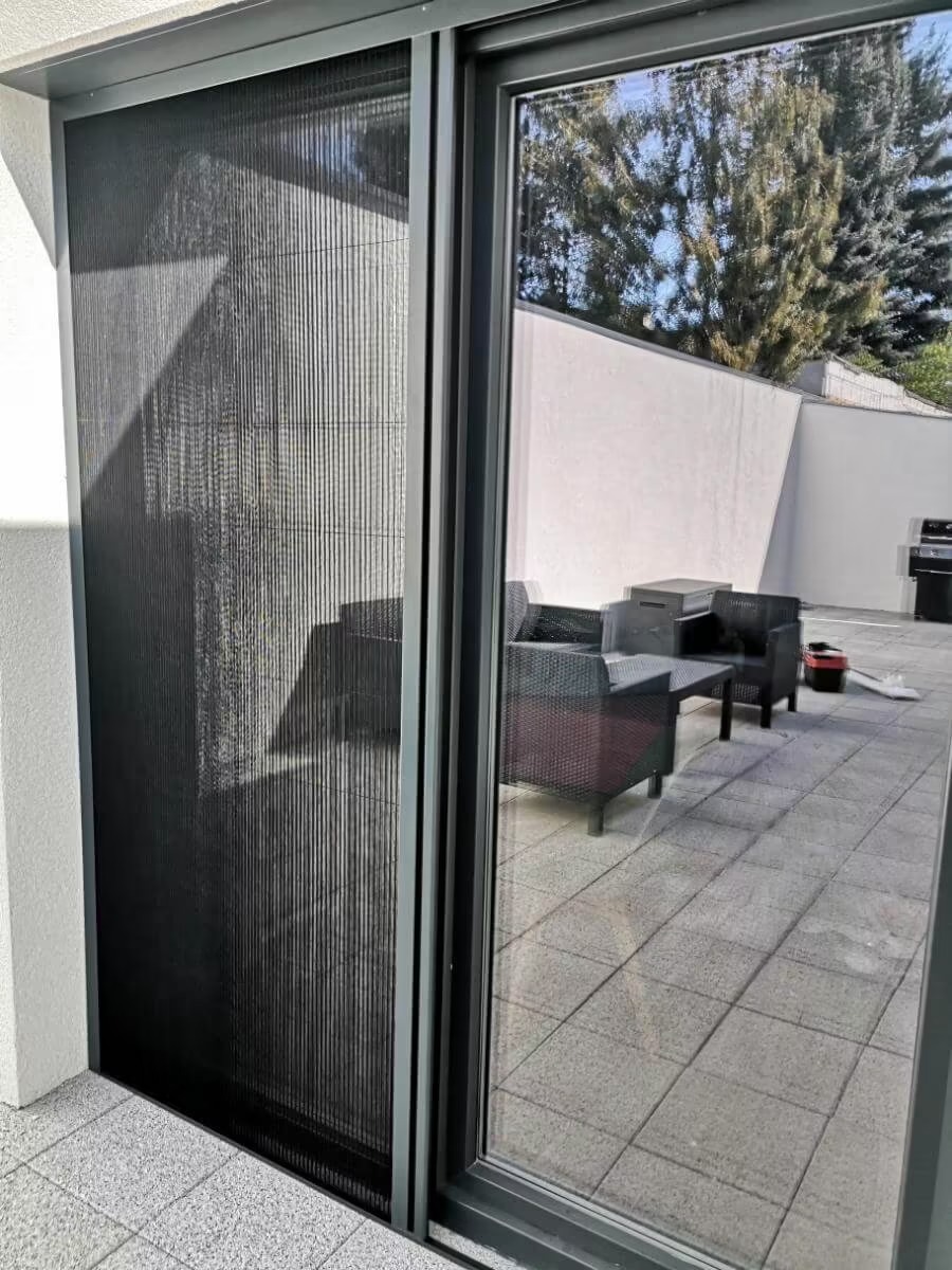 Zasłonięta plisowana moskitiera w szarej ramie, zamontowana na wyjściu na taras. Widoczny fragment tarasu z meblami ogrodowymi i drzewo w tle.