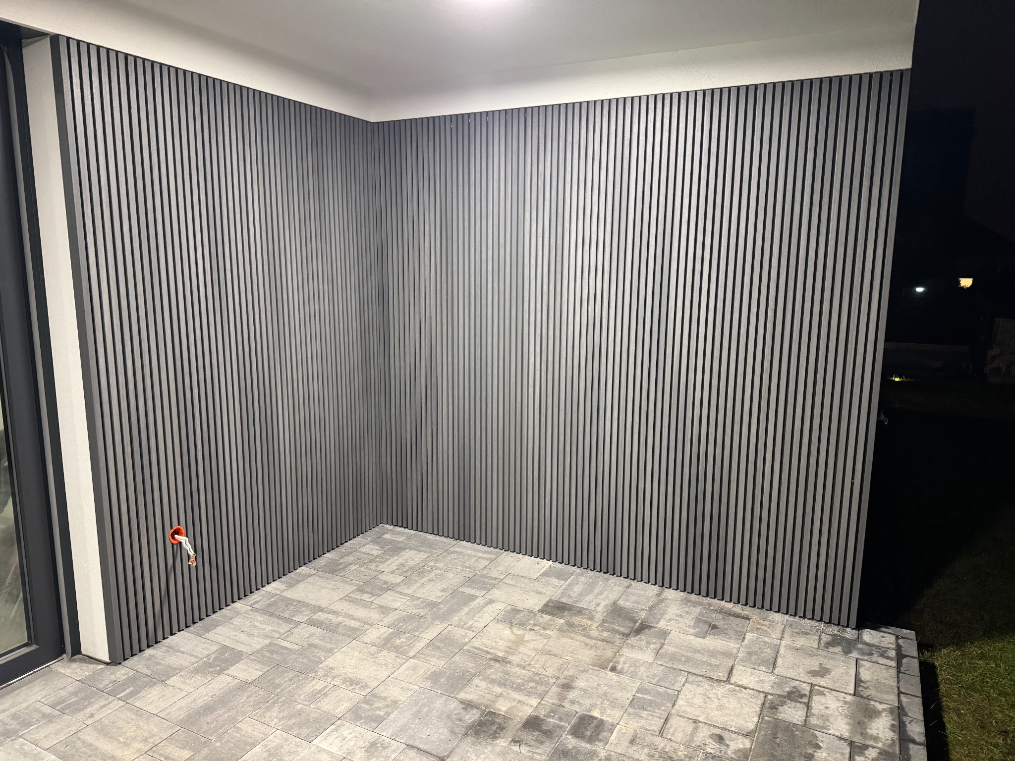 Nowoczesny taras z szarymi panelami ściennymi i kostką brukową. Widoczny fragment instalacji elektrycznej. Ujęcie z niskiego kąta, podkreślające geometrię.