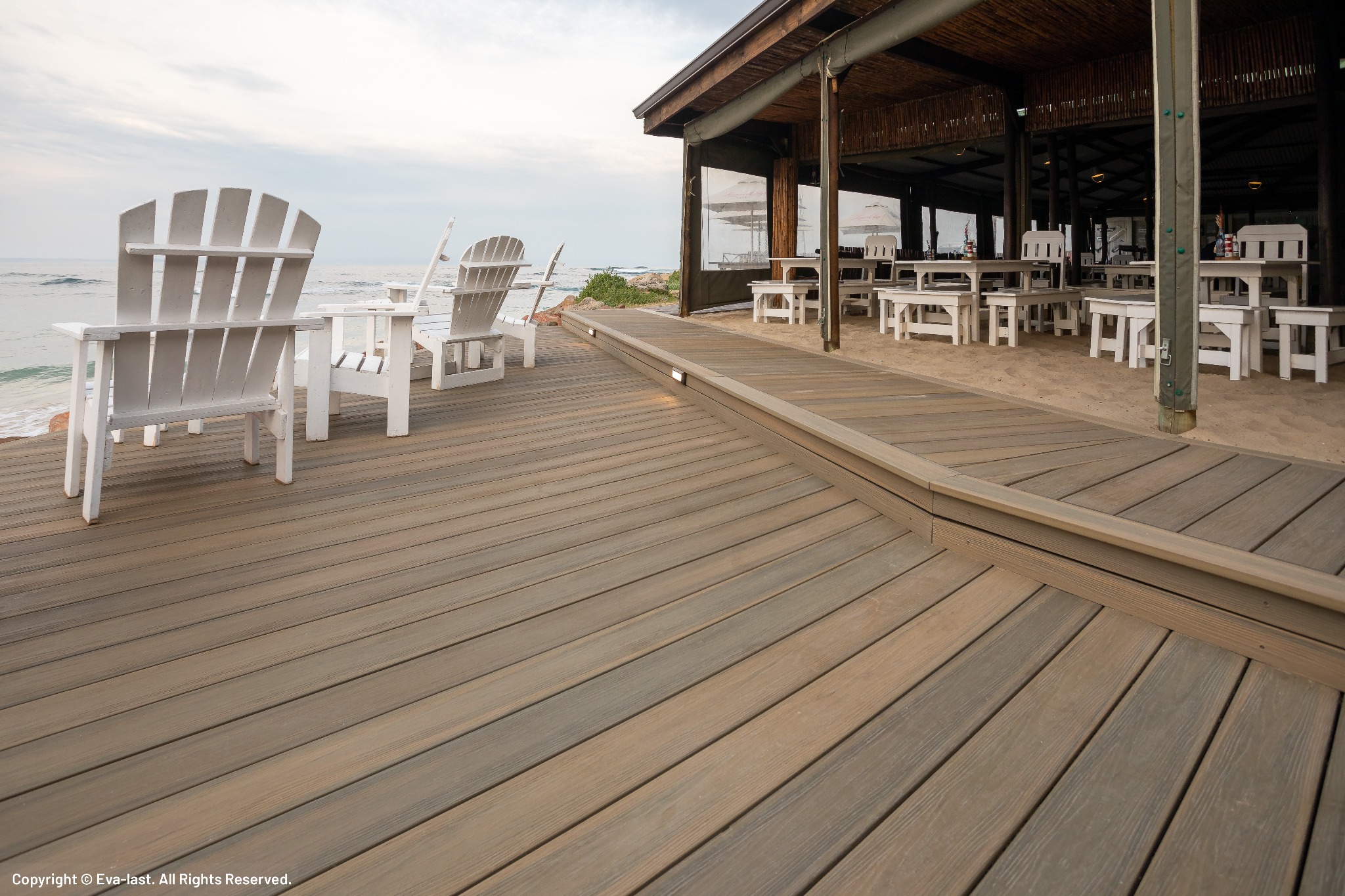 Kompozytowy taras z jasnego drewna prowadzi do restauracji na plaży. Białe krzesła Adirondack z widokiem na ocean. Oświetlenie wbudowane w podłogę tarasu.