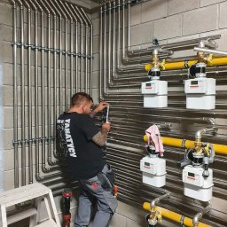 HYDROPEX SPÓŁKA Z OGRANICZONĄ ODPOWIEDZIALNOŚCIĄ - Instalacja gazowa w bloku z szarych cegieł. Hydraulik w czarnej koszulce mierzy rury. Widoczne liczniki gazu, żółte rury i narzędzia pracy.