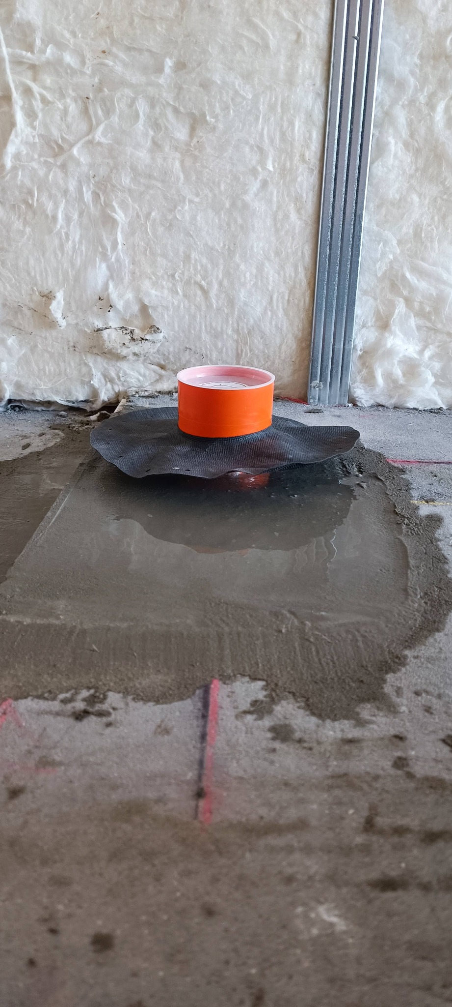 Pomarańczowy element instalacji na czarnej macie, zalany wodą na surowym betonie. W tle izolacja i metalowy profil. Widok z niskiej perspektywy.