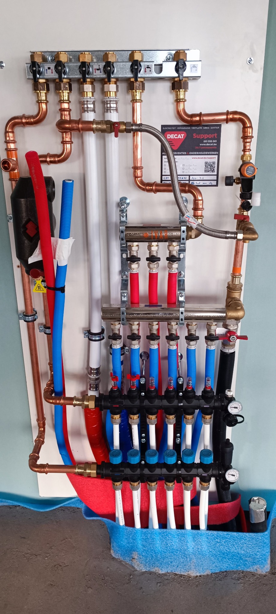 Rozdzielacz ogrzewania podłogowego z rurami miedzianymi, białymi, czerwonymi i niebieskimi, zamontowany na ścianie. Widoczne zawory i manometry.