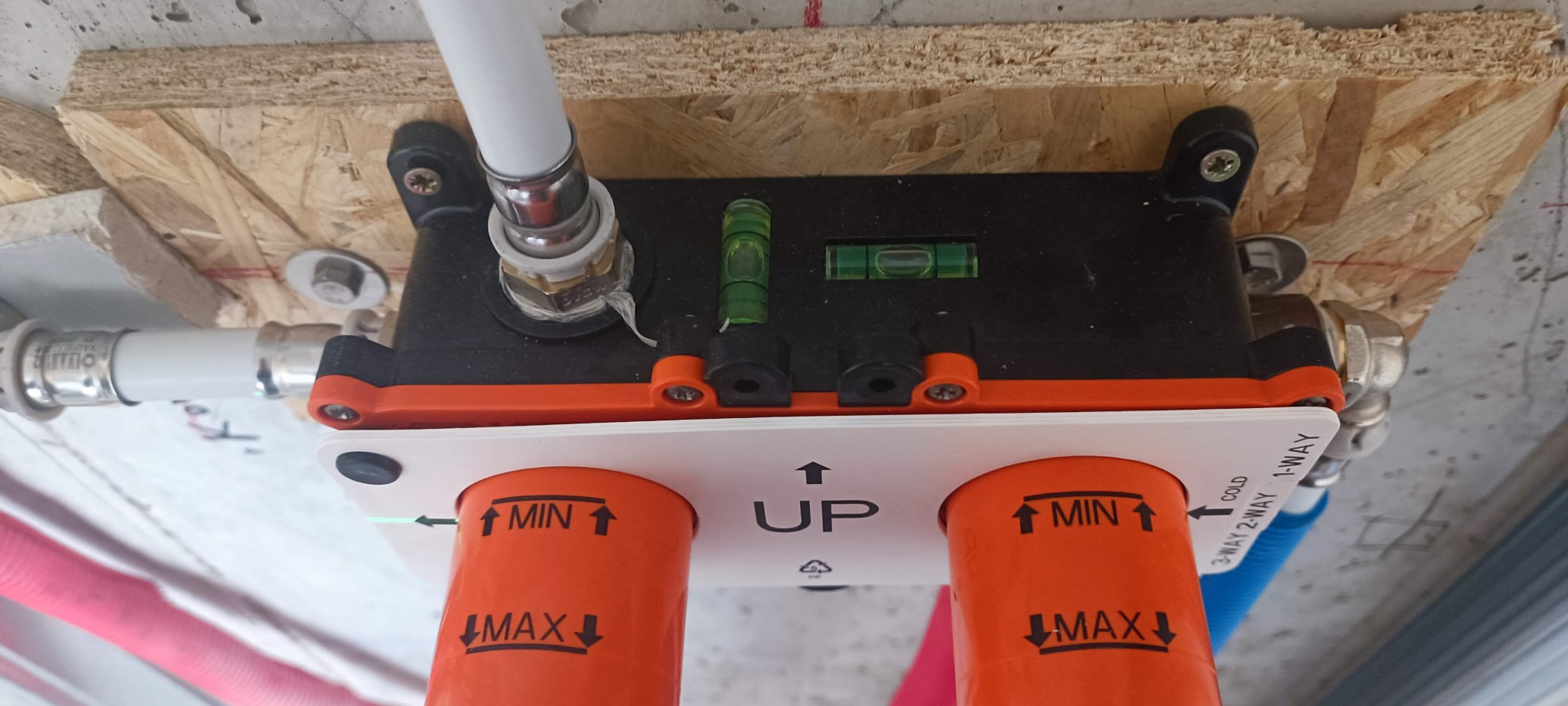 Rozdzielacz instalacji wodnej zamontowany do sufitu, z widocznymi rurami doprowadzającymi wodę. Poziomica wbudowana w rozdzielacz wskazuje prawidłowy montaż.