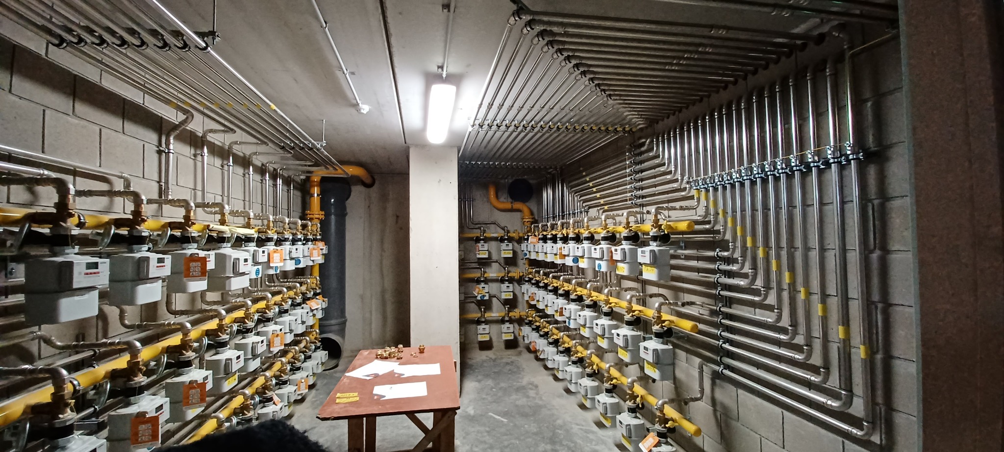 Węzeł gazowy z licznymi, równolegle ułożonymi rurami i gazomierzami na ścianach z betonu komórkowego. Na pierwszym planie stół z dokumentami i elementami instalacji.